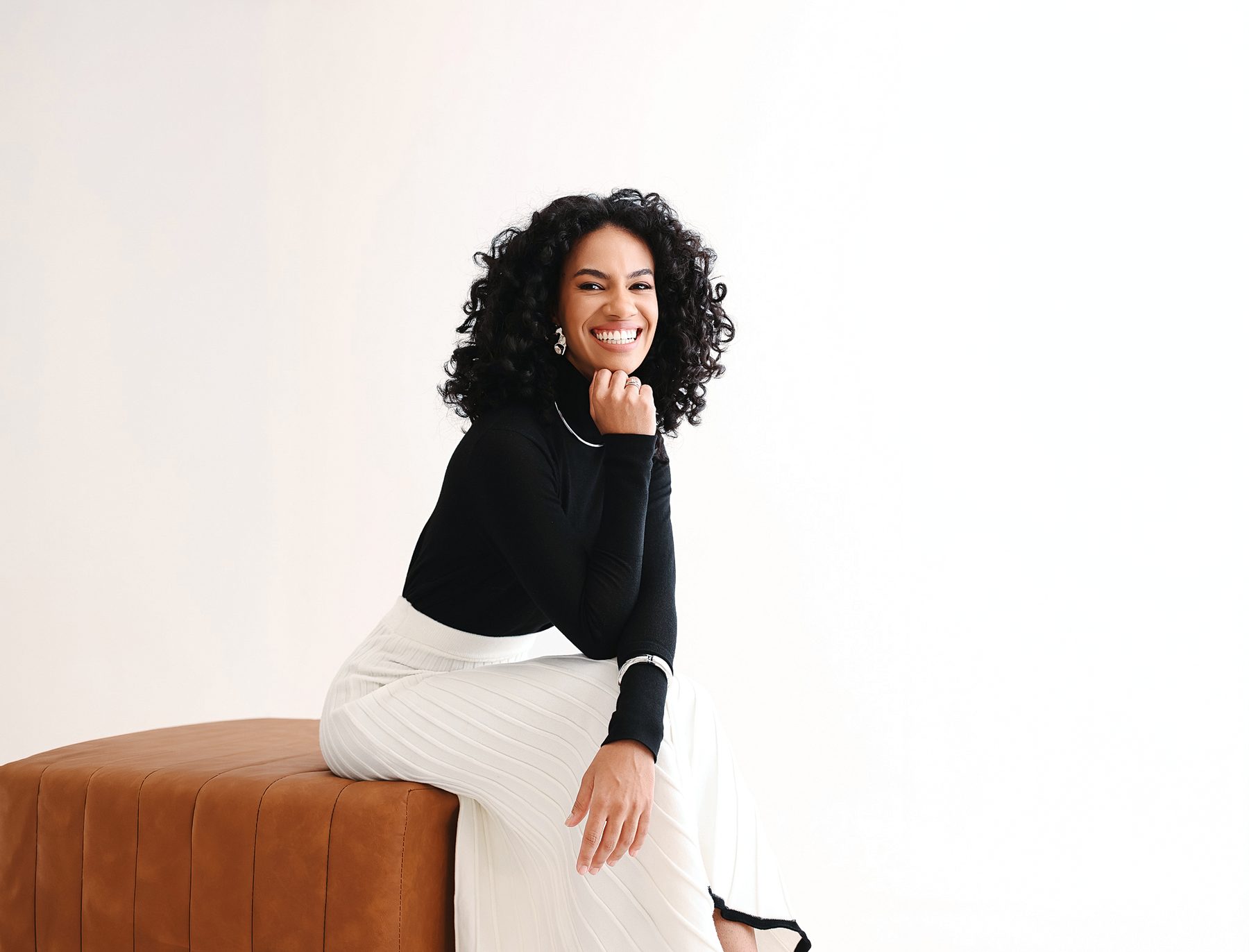 Dayane sentada, sorrindo, de gola alta preta e saia plissada branca